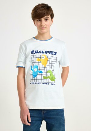 Chłopiec ubrany w biały t-shirt z grafiką przedstawiającą niebieskie, żółte i zielone postacie z kreskówek na kratkowanym tle, w połączeniu z niebieskimi dżinsami.