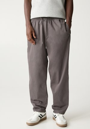 Homme portant un pantalon gris ample avec cordon de serrage et poches, ainsi que des baskets blanches à rayures noires sur un fond clair uni.