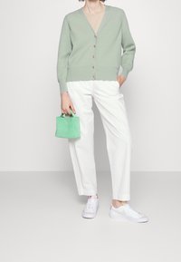 Cardigan verde menta con texture a coste e bottoni dorati, abbinato a pantaloni bianchi a gamba larga e sneakers bianche. Borsa verde piccola tenuta in mano.
