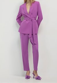 Fioletowa dopasowana marynarka z dekoltem w serek i paskiem w talii, zestawiona z pasującymi spodniami. Dodatki obejmują lawendowe szpilki z detalami w formie kokardek.