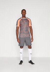 Graues Sportshirt mit orangen Akzenten und Wellenmuster, kombiniert mit grauen Shorts, weißen Kniestrümpfen und schwarzen Fußballschuhen.