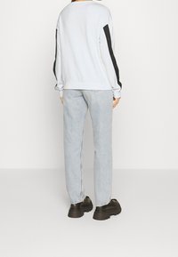 Persona con felpa bianca a maniche nere, jeans azzurri chiari e scarpe nere con suola spessa, in piedi davanti a uno sfondo bianco semplice.