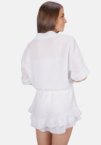 Robe en coton blanc avec un tissu texturé, manches courtes retroussées, col ouvert et détails de volants à l'ourlet. Vue de dos montrée.