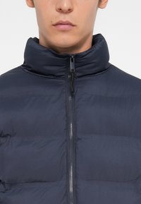 Marineblå puffjakke med høy krage, med glidelås foran og quiltet tekstur. Glatt stoff, lett design.