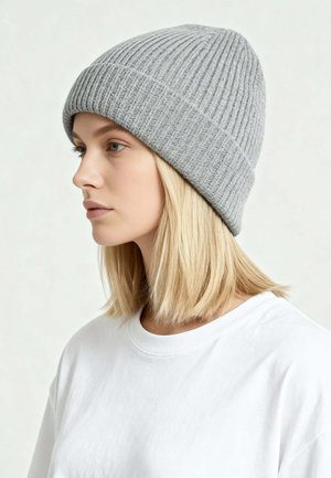 Jonge vrouw met blond haar, gekleed in een grijze geribbelde gebreide muts en een eenvoudig wit shirt, kijkt naar links tegen een neutrale achtergrond.