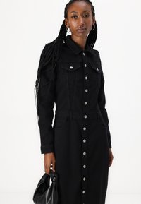 Robe en jean noir à manches longues, boutonnée sur le devant, avec deux poches poitrine, des poches latérales et une taille cintrée. Le mannequin tient un sac à main noir.