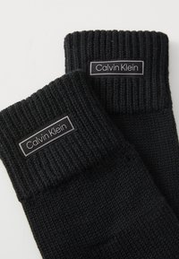 Svarte ankelsokker i strikket materiale med rillestruktur og foldet mansjett. Har en rektangulær "Calvin Klein"-etikett på hver sokk.