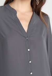 Blusa gris con escote en V, mangas largas y seis botones. Hecha de tela lisa; presenta detalles sutiles de costura.