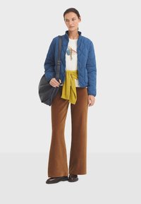 Quilted blue jacket, white t-shirt with graphic, yellow knot detail at waist, brown corduroy wide-leg pants, black bag, brown shoes.