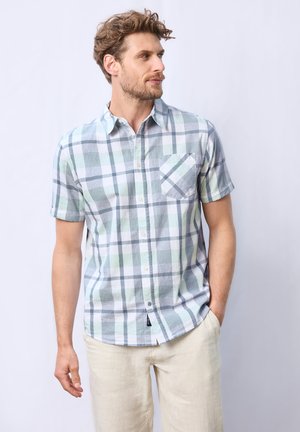 Hombre con cabello rizado que lleva una camisa de cuadros en tonos verde claro y gris de manga corta y pantalones beiges, mirando de lado con una mano en el bolsillo.