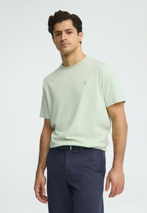 Uomo in piedi che indossa una maglietta a maniche corte verde chiaro e pantaloni blu scuro con cintura, su sfondo grigio uniforme.