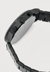 Μαύρο μεταλλικό ρολόι χειρός με συνδεδεμένο λουράκι, που διαθέτει στρογγυλή θήκη και τρία κουμπιά στα πλάγια, φωτογραφημένο σε απλή ανοιχτόχρωμη φόντο.