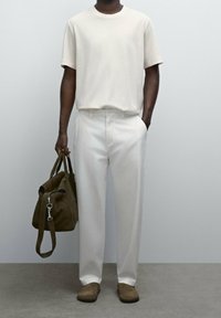 Man i vit t-shirt och byxor, som håller en olivgrön weekendväska i ena handen, stående på en grå golv mot en slät vägg.