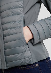 Giacca grigia con texture trapuntata e gonfia sulla manica sinistra, tessuto liscio sul corpo e tasca laterale con dettaglio della cerniera.