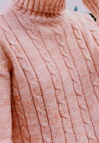 Gros plan d'un pull en maille torsadée rose pâle avec un col roulé épais côtelé et des motifs tressés texturés.