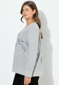 Sudadera gris de manga larga, con un corte relajado y un diseño gráfico que presenta un patrón estilizado en la parte frontal. Textura suave.