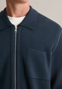 Granatowa kurtka zapinana na zamek z teksturowanym kołnierzem, ściągaczami na mankietach, subtelnym logo na kieszeni oraz kontrastującym przeszyciem na ramieniu.
