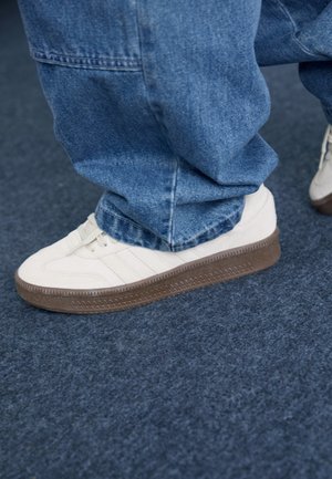White sneakers with textured brown rubber soles, featuring three horizontal stripes and a smooth upper on denim pants.