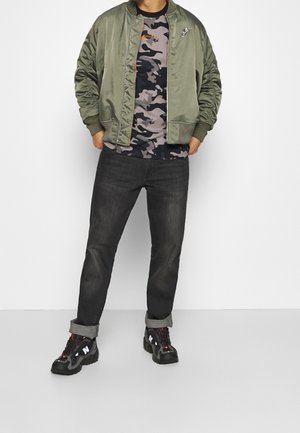 Olijfgroene bomberjack, grijs camo T-shirt, zwarte jeans met opgerolde manchetten, en stevige zwarte sneakers met rode accenten.