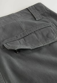 Gesäßtasche von dunkelgrauen Hosen mit Klappe, die mit einem schwarzen Druckknopf gesichert ist, und einem Gürtelhalfter über der Tasche.