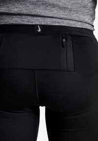 Primo piano di leggings athletic neri con tasca posteriore con zip e cintura con un piccolo logo riflettente Nike, indossati con una maglietta grigia.