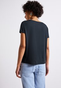 Haut noir à manches courtes, à coupe décontractée, texture lisse, encolure arrondie, porté avec un jean en denim bleu clair, design visible au dos.