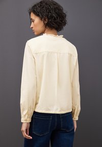 Blusa beige chiaro con scollatura arricciata e polsini elasticizzati, caratterizzata da un design ampio e una piega sul retro per una maggiore comodità.