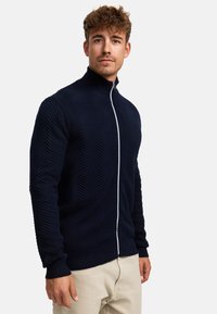 Marineblå zip-up genser med et teksturert herringbone-mønster, høy krage og hvit glidelås, parret med lyse bukser.