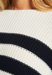 Gros plan d'un pull tricoté avec de larges rayures horizontales noires et blanches porté par une personne aux longs cheveux blonds.