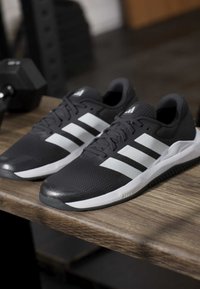 Paire de chaussures de sport noires avec des bandes blanches et des semelles posées sur une surface en bois avec un haltère en arrière-plan.