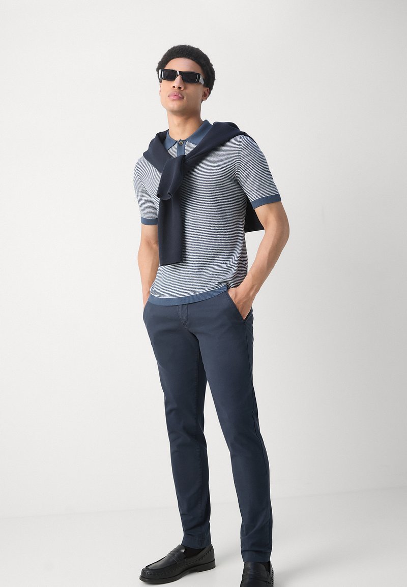 Hellblau-weiß gestreiftes kurzärmliges Poloshirt, navyfarbene Hose, schwarze Schuhe und Sonnenbrille, mit einem navyfarbigen Pullover lässig über die Schultern gelegt.
