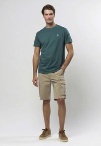 Tirkizno plava majica s okruglim izrezom i malim logotipom, uparen s bež cargo kratkim hlačama sa bočnim džepovima i bež smeđim tenisicama od brušene kože. Neutralna pozadina.