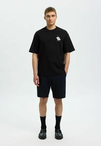 Juodas medvilninis T-shirt su trumpomis rankovėmis ir mažais grafikos elementais. Derinamas su tamsiai mėlynomis tekstūruotomis šortais ir juodomis kojinėmis bei batais.