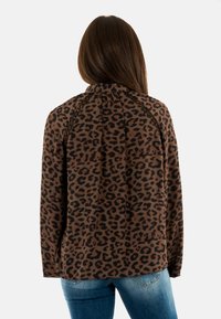 Donna con lunghi capelli castani che indossa una blusa a stampa leopardo marrone e nera e jeans blu, di spalle su uno sfondo bianco.