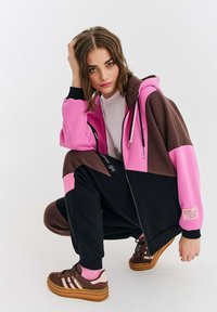 Młoda kobieta w squacie, ubrana w różową, brązową i czarną bluzę z zamkiem, dopasowane spodnie, różowe skarpetki oraz brązowe trampki Adidas Gazelle.