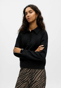 Schwarzer Pullover mit Kragen, Knopfleiste und Raglanärmel, kombiniert mit braunen und schwarzen gestreiften Hosen im Zebra-Print.