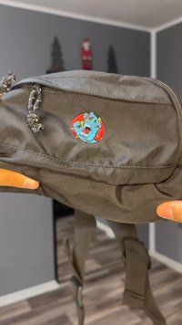 Grønn nylon midjeveske med glidelåslomme, med et fargerikt brodert merke av en hånd på en rød sirkel. Justerbar stropp synlig.