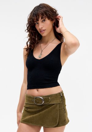 Femme aux cheveux bouclés portant un crop top noir à motifs et une mini-jupe en velours côtelé vert olive avec une grande boucle de ceinture circulaire.