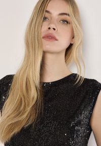 Jonge vrouw met lang blond haar, gekleed in een zwarte mouwloze top met pailletten, tegen een effen lichte achtergrond.