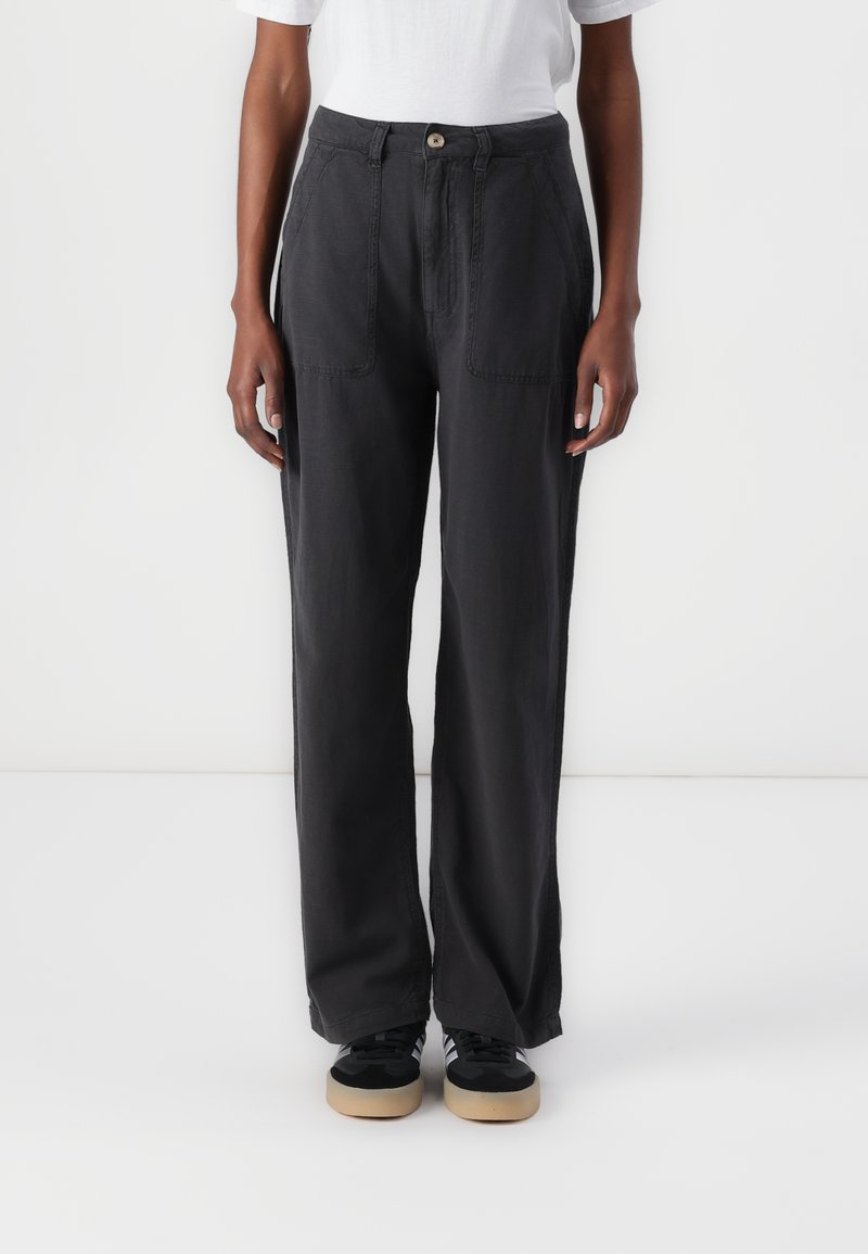 Pantalons larges noirs en tissu doux avec deux poches avant, une fermeture à bouton et une coupe ample, portés avec des baskets noires.