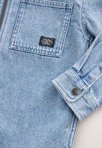 Light blue denim jacket featuring a chest pocket with a logo patch, silver snap button closure on the cuff, and a classic fit design.