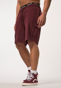 Pantalones cortos burdeos con cintura con cordón, bolsillo lateral y banda elástica que presenta "STHUGE". Llevados con calcetines blancos y zapatillas burdeos.
