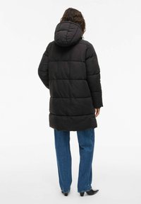 Persona, kas ģērbjas garā melnā pūkainajā mētelī ar kapuci, zilos džinsos un melnās norādējuma kurpēs, stāv ar muguru pret neitrālu baltu fonu.