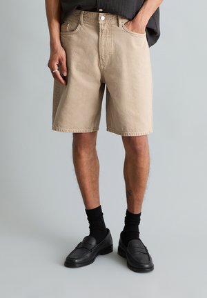 Persoon draagt beige knielange shorts, zwarte sokken en zwarte loafers, staat met één hand in de zak tegen een effen achtergrond.
