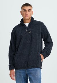 Pullover in pile blu scuro con colletto alto, zip parziale frontale e tasche laterali. Superficie morbida e texturizzata con un piccolo etichetta del marchio sul petto.