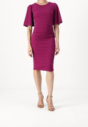 Frau trägt ein figurbetontes, knielanges magentafarbenes Kleid mit weiten kurzen Ärmeln und hautfarbene offene Sandalen mit Knöchelriemen, stehend vor weißem Hintergrund.