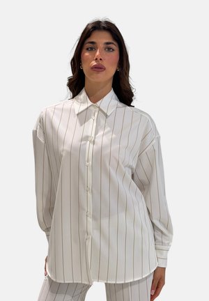 Donna che indossa una camicia bianca a maniche lunghe con bottoni e pantaloni abbinati con sottili strisce verticali, in piedi davanti a uno sfondo semplice.