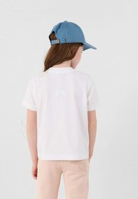 Witte katoenen T-shirt met korte mouwen, met een klein logo op de achterkant. Gecombineerd met lichtroze sweatpants en een blauwe pet.