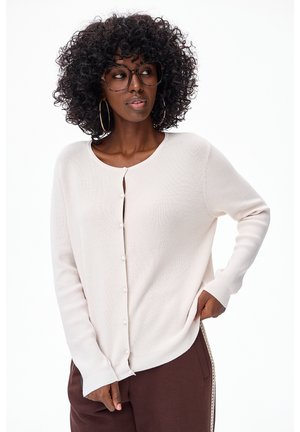 Vrouw met krullend haar, grote bril, gouden oorringen, een wit vest met knopen en bruine broek met zijstrepen, poserend met haar hand in haar zak.