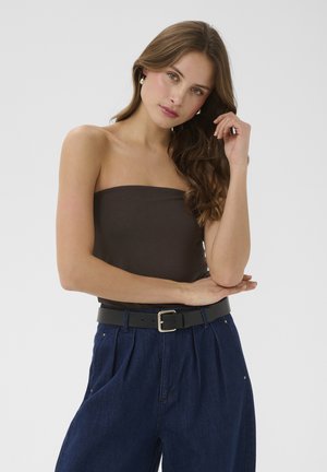 Tânără cu păr lung și bălai, purtând un top negru fără bretele, blugi albaștri cu talie înaltă, curea neagră și cercei de perlă, pozând pe un fundal alb.